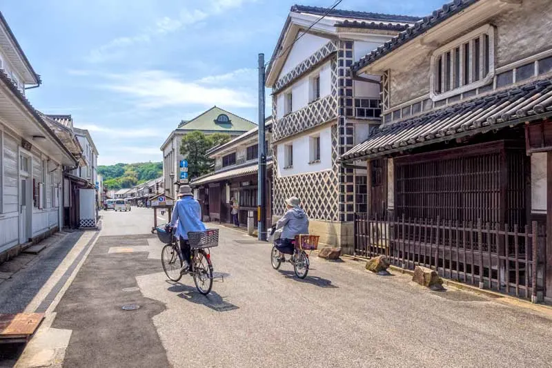 A traditional street in Kurashiki Bikan with local cyclists passing one of the old whte walled rice storehouses